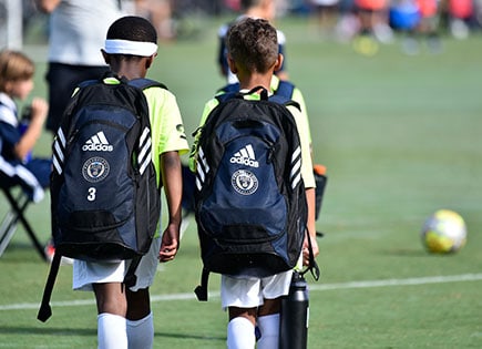 Two young children walking on a soccer field with blue adidas backpacks on.